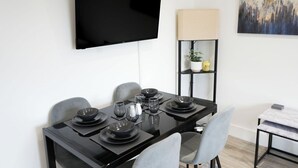 Un espace repas moderne avec une table noire dressée pour six personnes, un téléviseur à écran plat fixé au mur et une étagère minimaliste agrémentée d’une lampe et d’objets décoratifs.