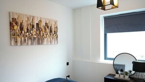 Une chambre à coucher avec un lit, une commode, un miroir, une fenêtre et un tableau représentant un paysage urbain accroché au mur.