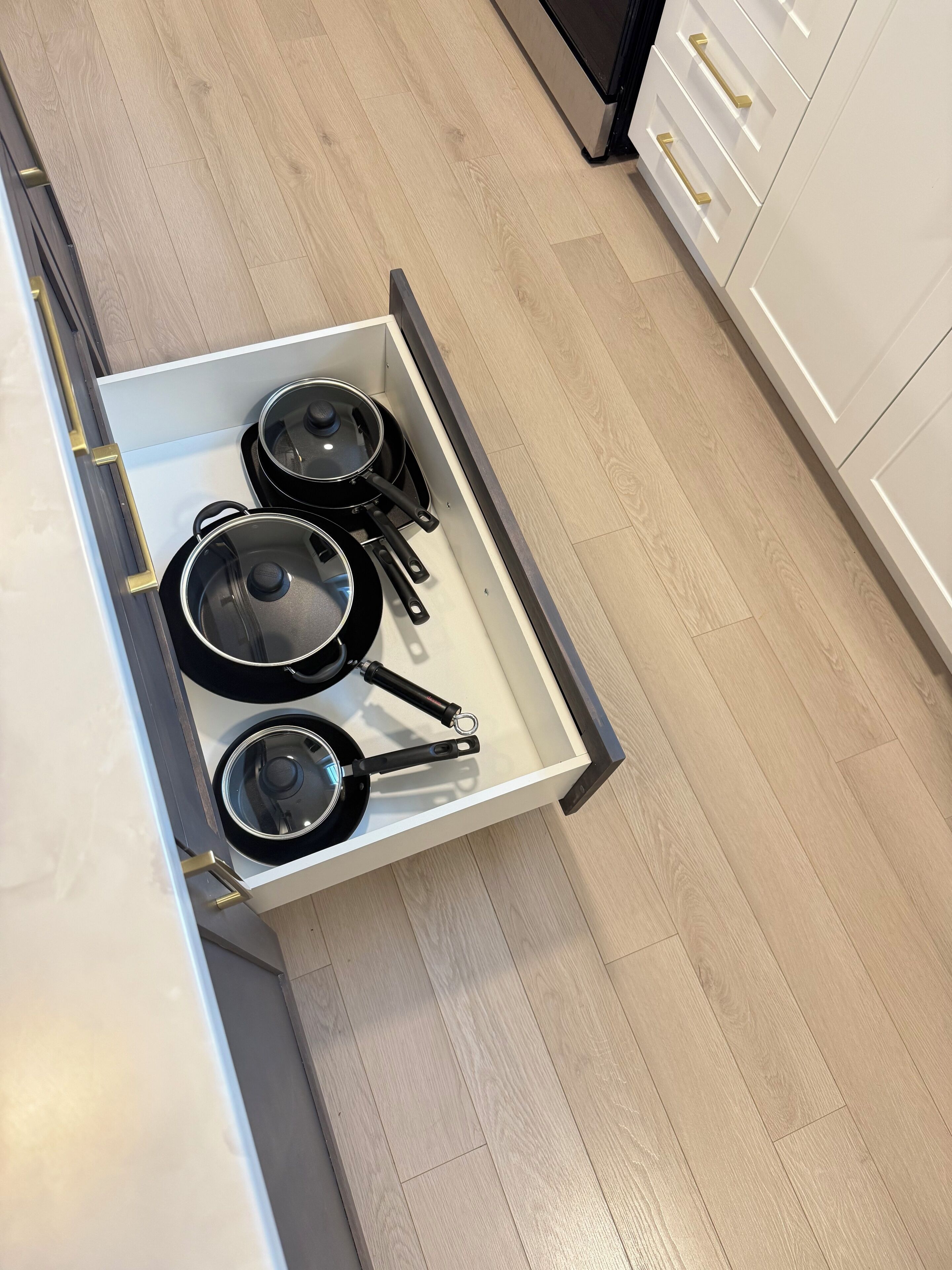 Une cuisine avec des meubles blancs, un tiroir contenant des casseroles et des poêles, et un plancher en bois.
