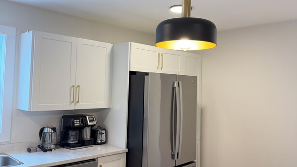 Una cocina moderna con gabinetes blancos, un refrigerador de acero inoxidable, una cafetera y una lámpara colgante.