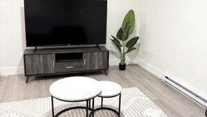 Una sala de estar con un televisor de pantalla plana, un mueble de madera oscura para el equipo de sonido, dos mesas redondas blancas, un tapete estampado y una planta verde grande.