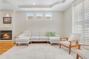Un salon comprenant un canapé d’angle, un fauteuil en bois, une cheminée et trois fenêtres donnant sur un beau paysage.