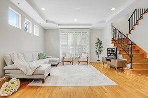 Un salon comprenant un canapé, des fauteuils, un téléviseur, un escalier et une grande fenêtre avec des stores.
