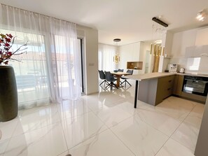 A modern kitchen with a dining area, a large window with sheer curtains, and a potted plant.