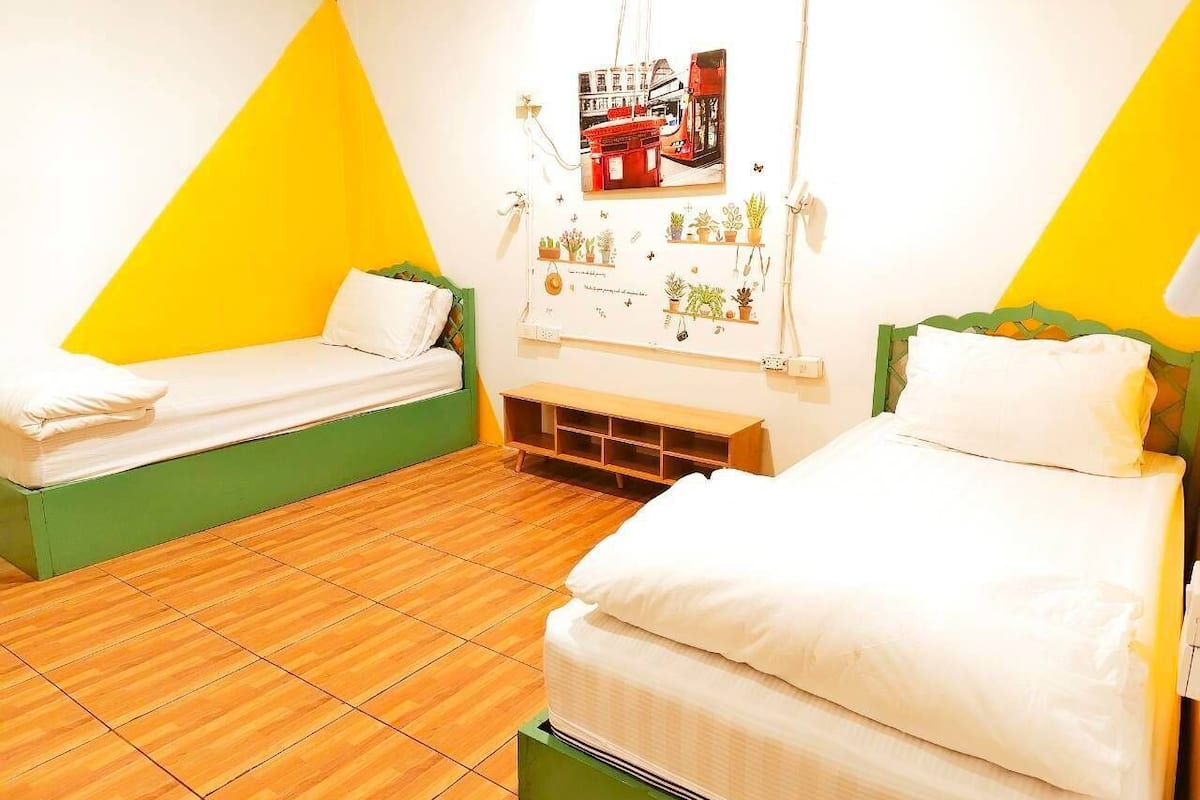 Quarto com duas camas, piso de madeira e parede com um quadro e pequenas plantas em vasos.