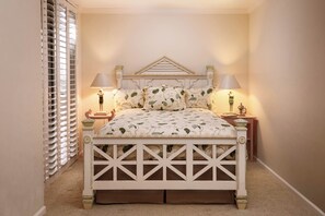 Quarto com cama, duas mesas de cabeceira, um criado-mudo, janela com persianas e piso coberto por carpete.