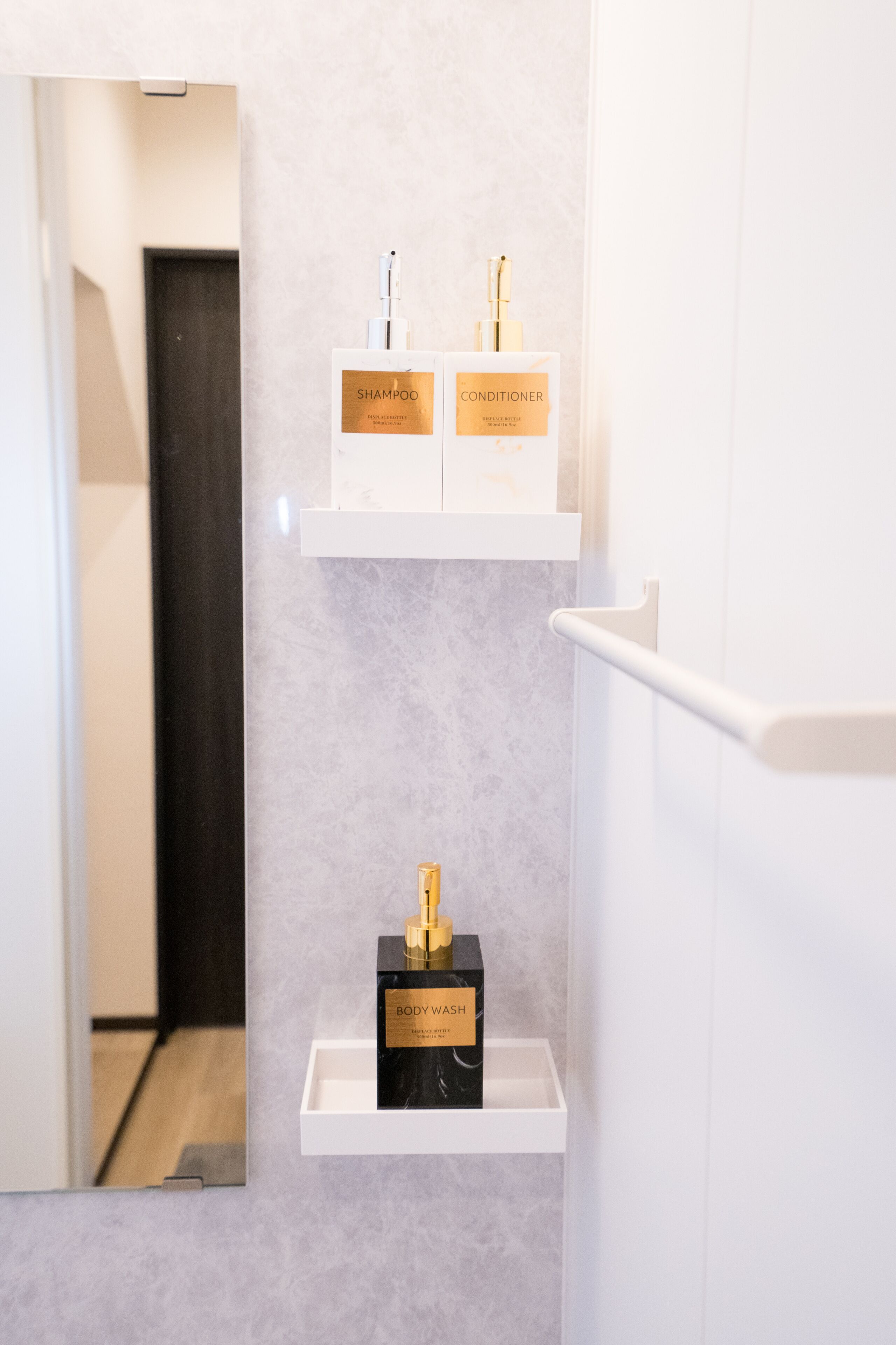 Et badeværelse med et spejl, to hvide hylder og tre flasker mærket 'SHAMPOO', 'CONDITIONER' og 'BODY WASH'.