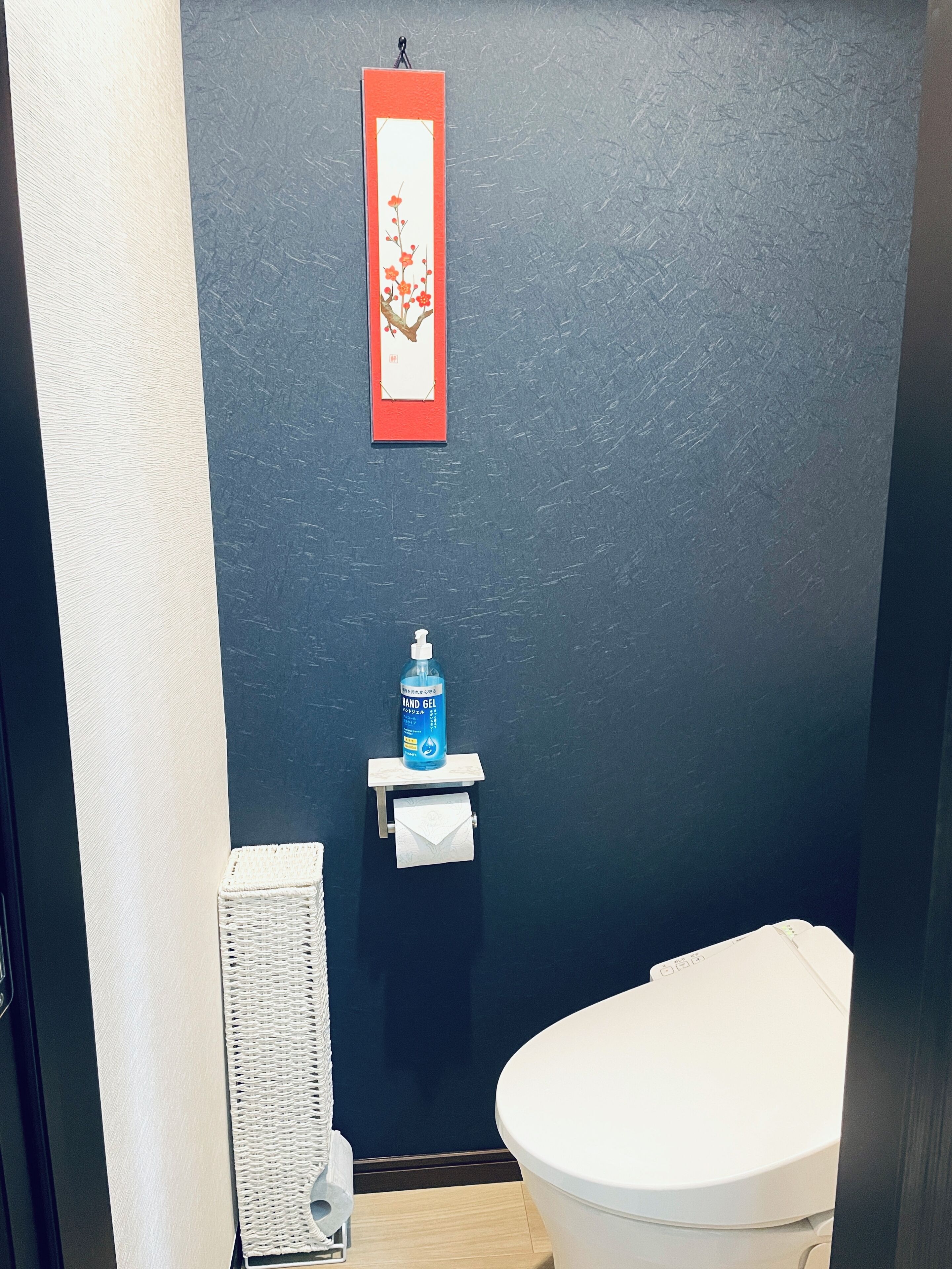 A small bathroom with a white toilet, a hand soap dispenser on a small shelf, a white towel hanging on a hook, and a framed picture on the wall.