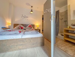Ein Schlafzimmer mit Bett, Nachttischen, einer Wandleuchte, einer Duschkabine und einem Badezimmer mit Waschbecken und Spiegel.