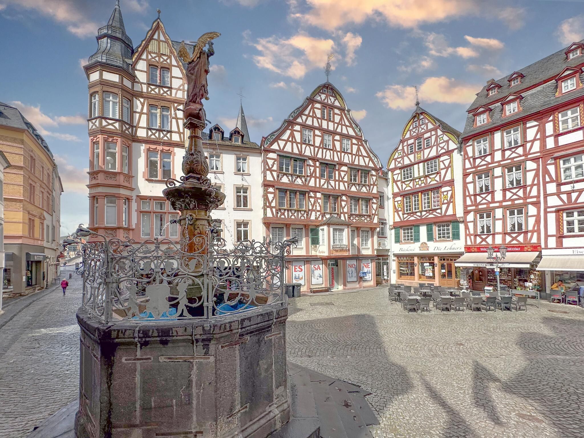 Une place pavée, bordée de bâtiments traditionnels à colombages, avec une fontaine et des tables installées à l’extérieur.