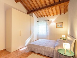 Ein Schlafzimmer mit einem Bett, einem Kleiderschrank, einem Fenster mit Vorhängen, einem Nachttisch mit Lampe und einem gerahmten Bild an der Wand.