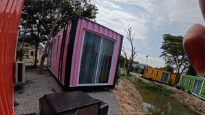 Un edificio tipo contenedor de color rosa y negro con ventanas amplias, una pequeña mesa con bancos y un estanque al fondo.