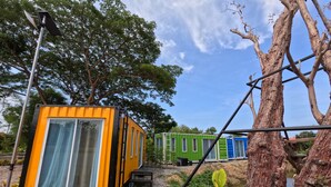 Una hilera de cabañas modulares coloridas con ventanas amplias, ubicada en un camino de grava con árboles al fondo.