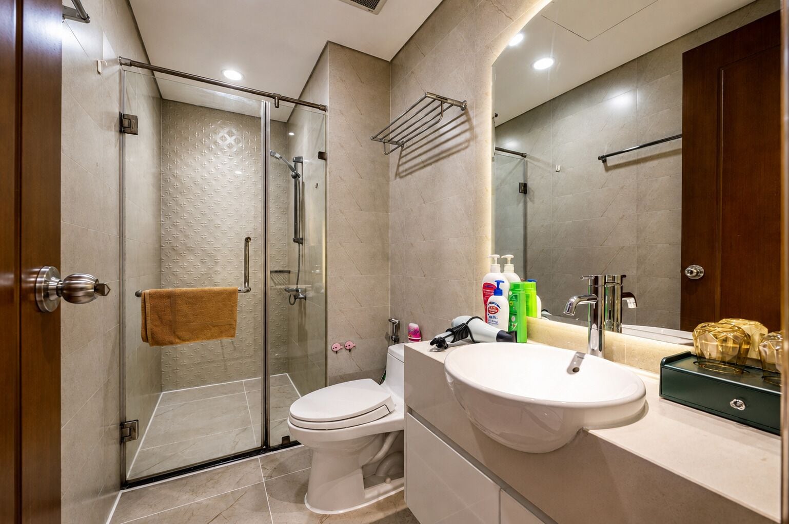 A modern bathroom with a glass-enclosed shower, a toilet, a sink with toiletries, and a large mirror.