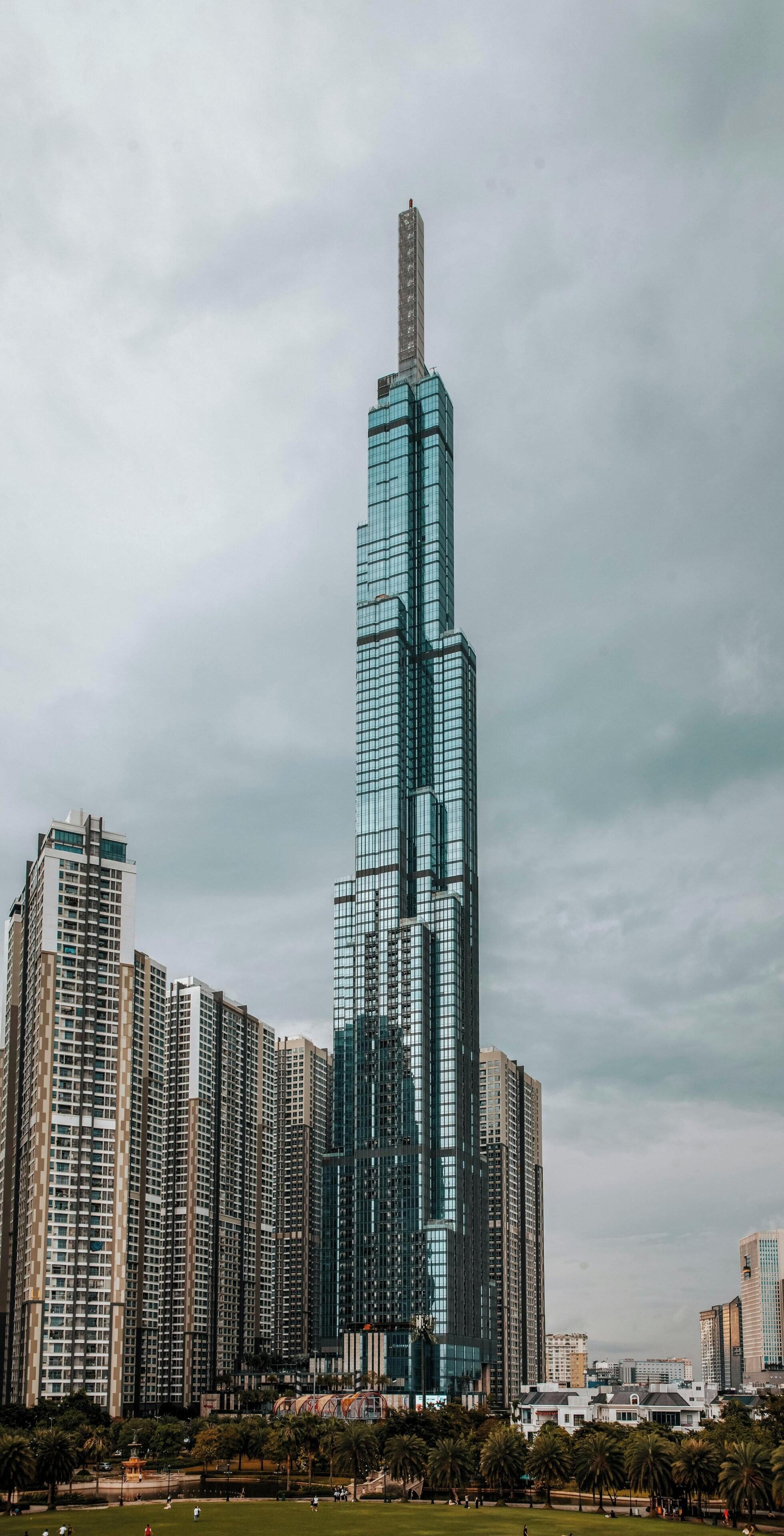 Un edificio de gran altura con fachada de vidrio, rodeado de otros edificios y un parque con palmeras.