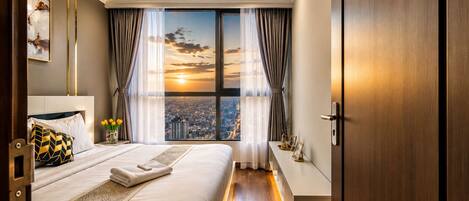 A hotel room with a large bed, a city view through the window, and a wooden floor.