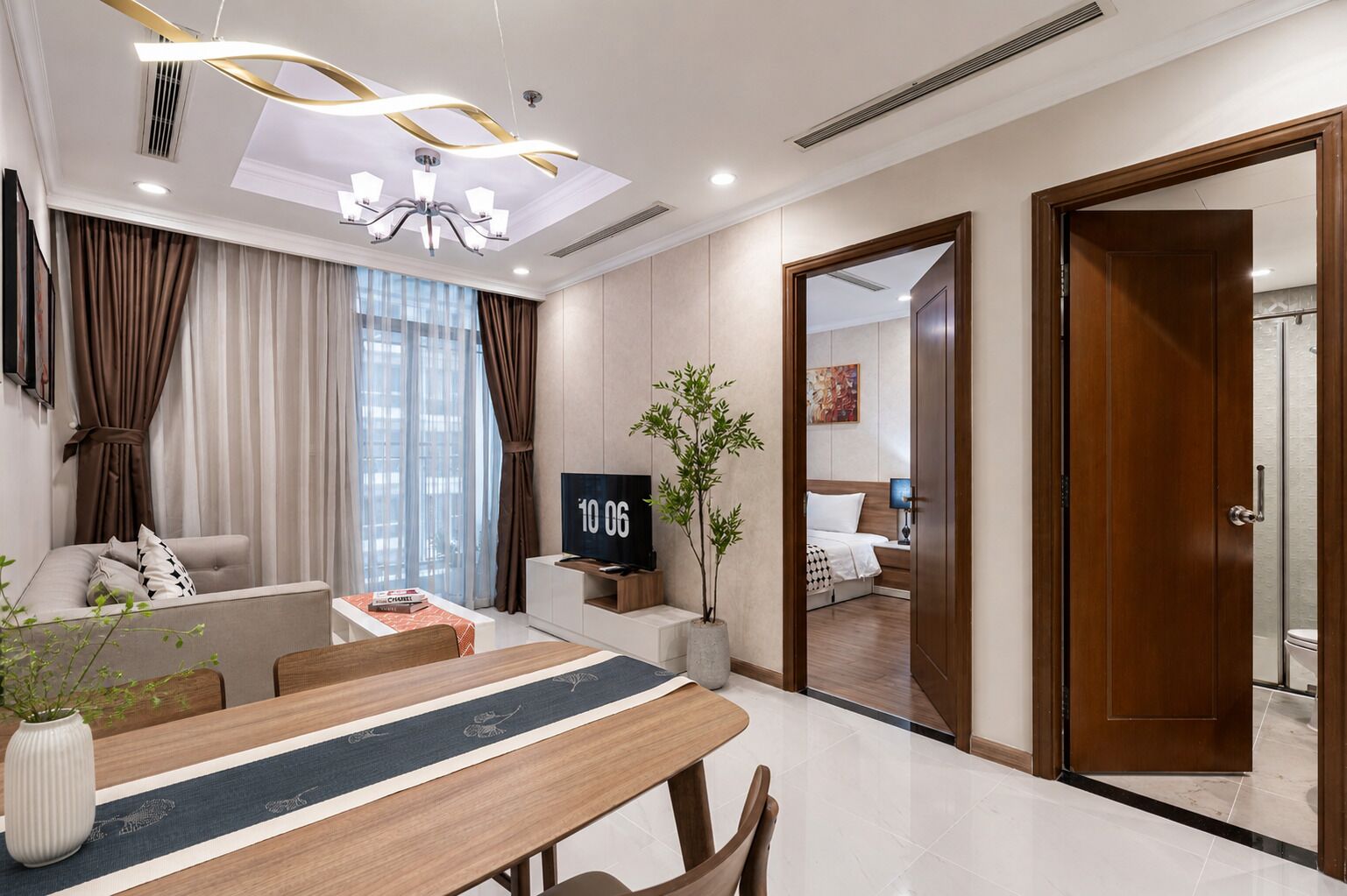 A modern hotel room with a dining area, a television, a plant, and a view of the adjacent bedroom.