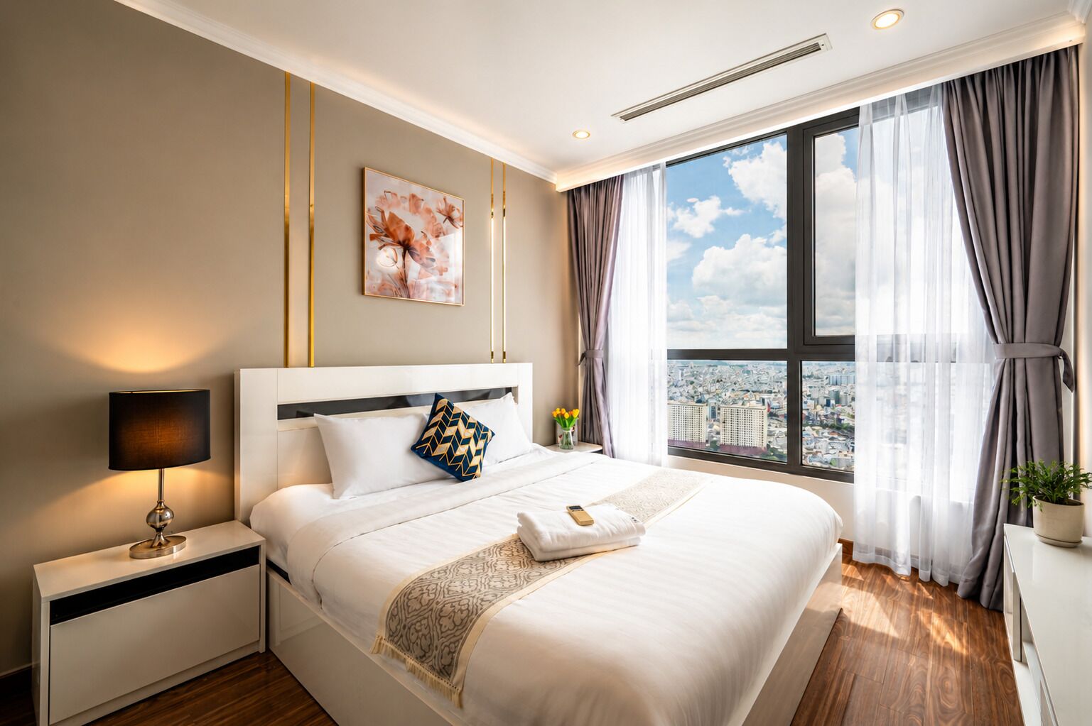 A hotel room with a large bed, a bedside table with a lamp, a view of the city through the window, and a painting on the wall.