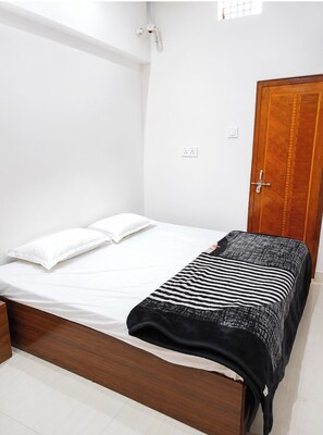 A bedroom with a wooden bed frame, white bedding, a black and white striped blanket, a wooden wardrobe, and a small window. - Hotel Ramesht Home Stay Near Kashi Ghat  (Varanasi)