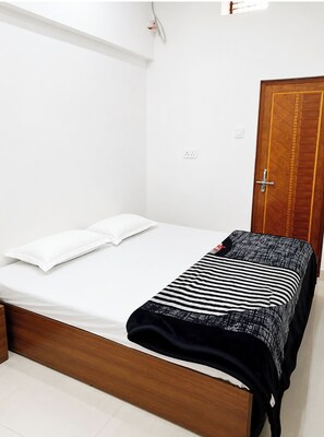 A single bed with white bedding, a wooden headboard, a black and white striped blanket, and a wooden door in a room with white walls and a tiled floor. - Hotel Ramesht Home Stay Near Kashi Ghat  (Varanasi)