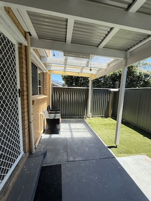 A covered patio area with a seating arrangement, a metal gate, and a fenced backyard. - Beach Flat 2, Surfside on Bribie (hear the surf) (Woorim)
