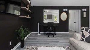 A modern living room with a black wall, a flat-screen TV, a desk with a chair, a round mirror, and a sofa with a patterned cushion. - South KC upscale Corporate Rental (Grandview)