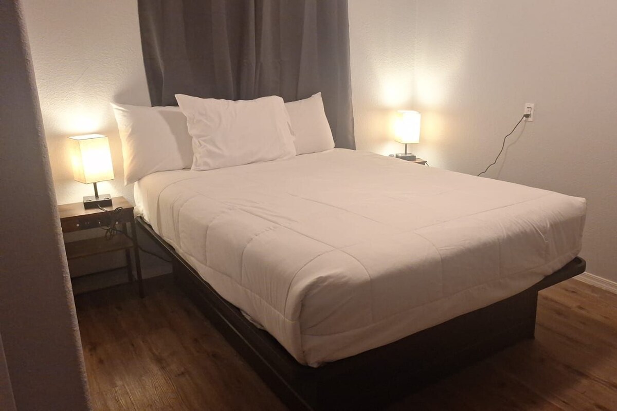 A neatly made bed with a white comforter and pillows, two bedside lamps, a wooden nightstand, and a power outlet on the wall.