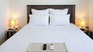 A hotel room with a bed, two bedside lamps, a ceiling fan, an air conditioner, and a bedside table with a book and a glass. - Grand Canyon Economy Rentals (Williams)