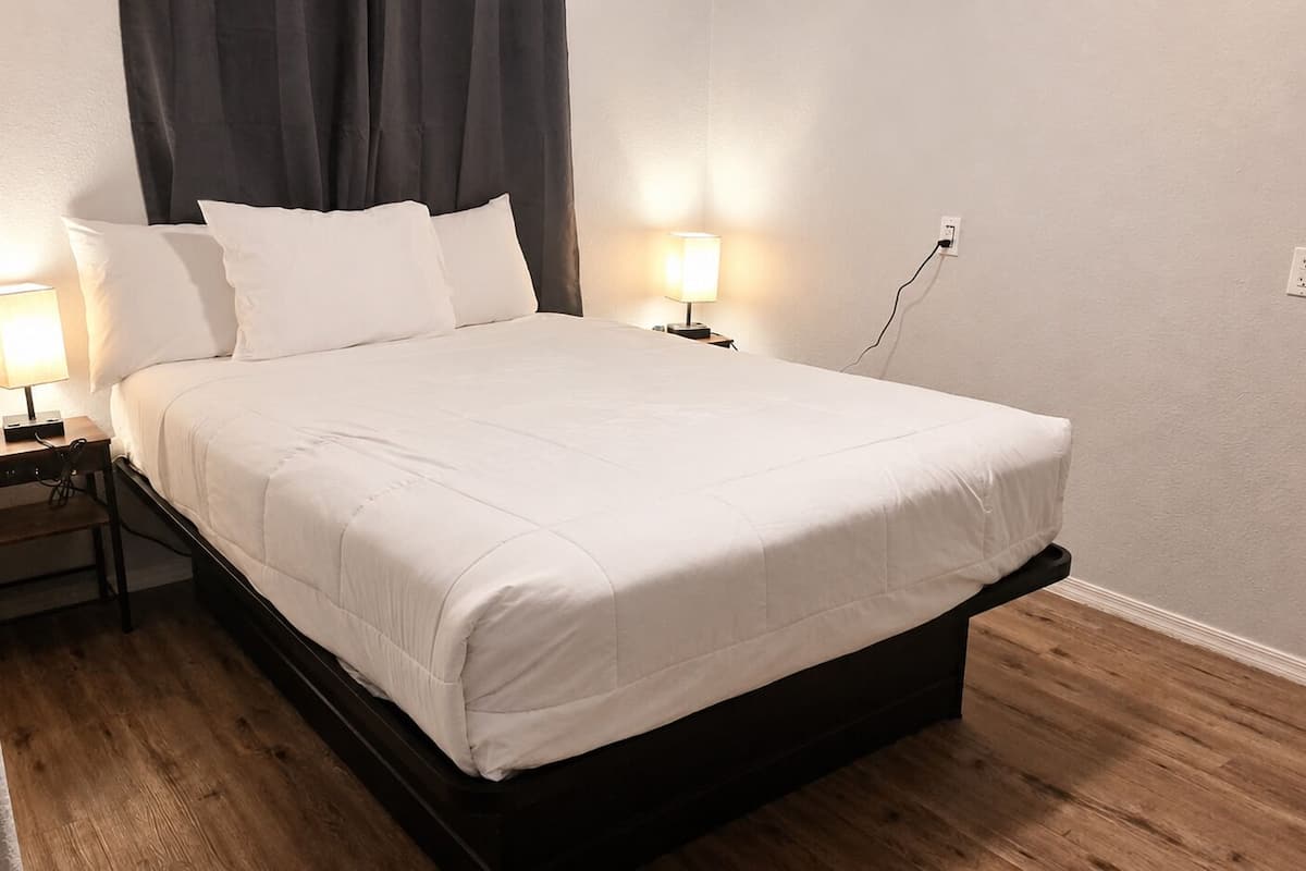 Een netjes opgemaakt bed met witte lakens, twee nachtkastjes met lampen, een donker hoofdbord en een houten vloer.