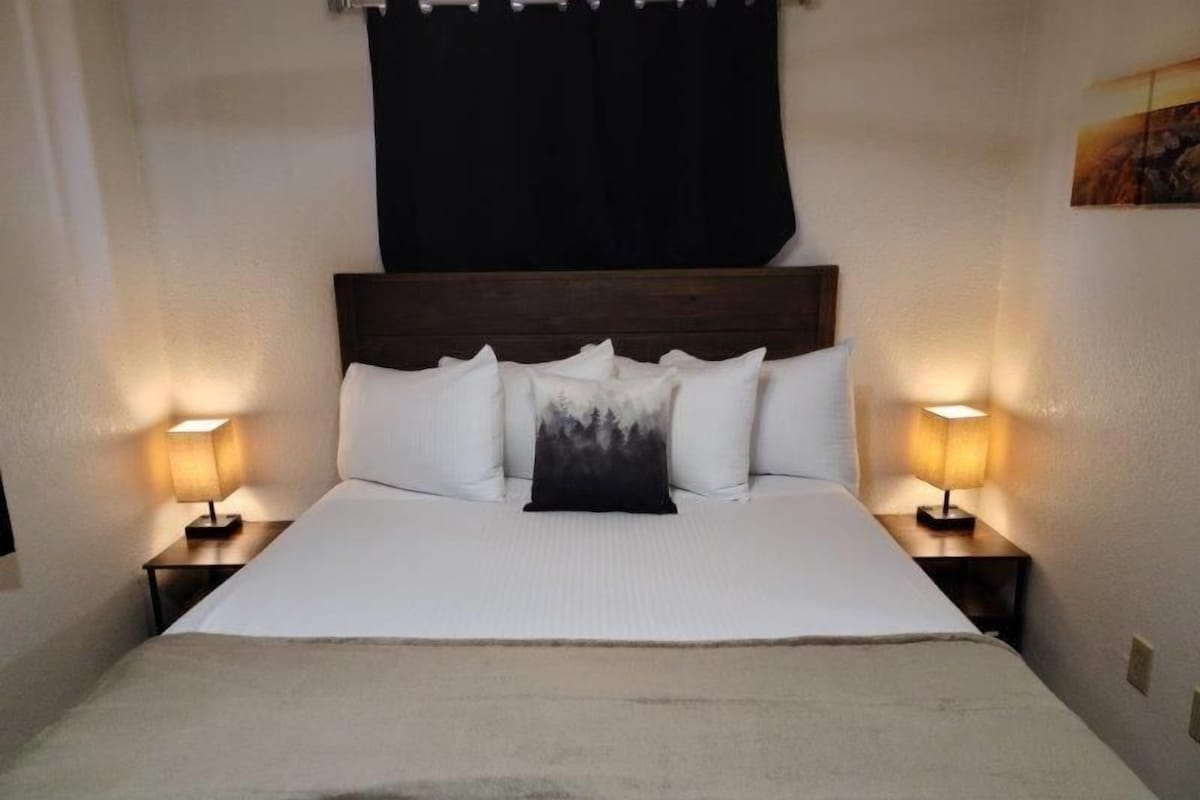 A hotel room with a large bed, two bedside tables with lamps, a dark headboard, and a painting on the wall.