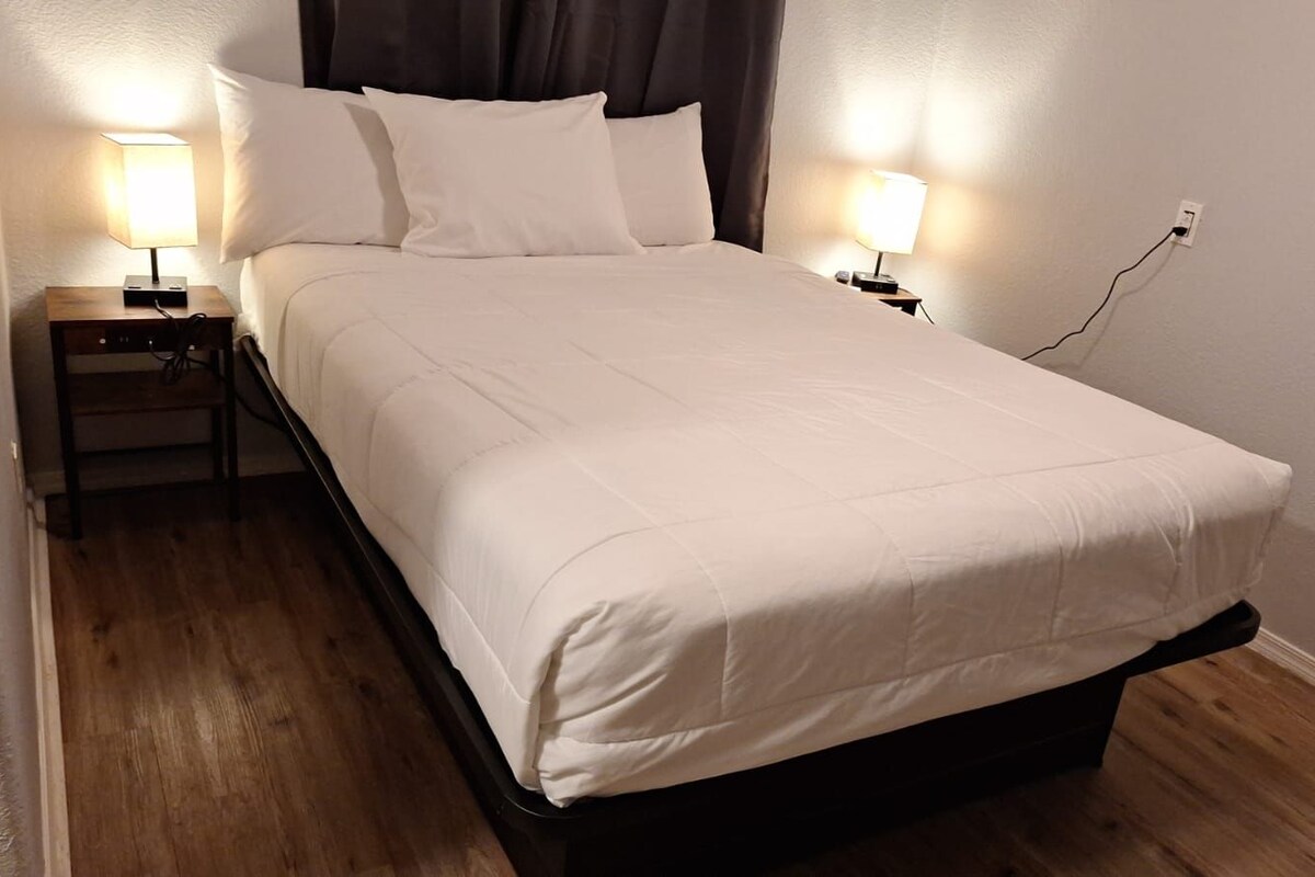 A neatly made bed with a white duvet, two bedside lamps, and a power outlet on the wall.