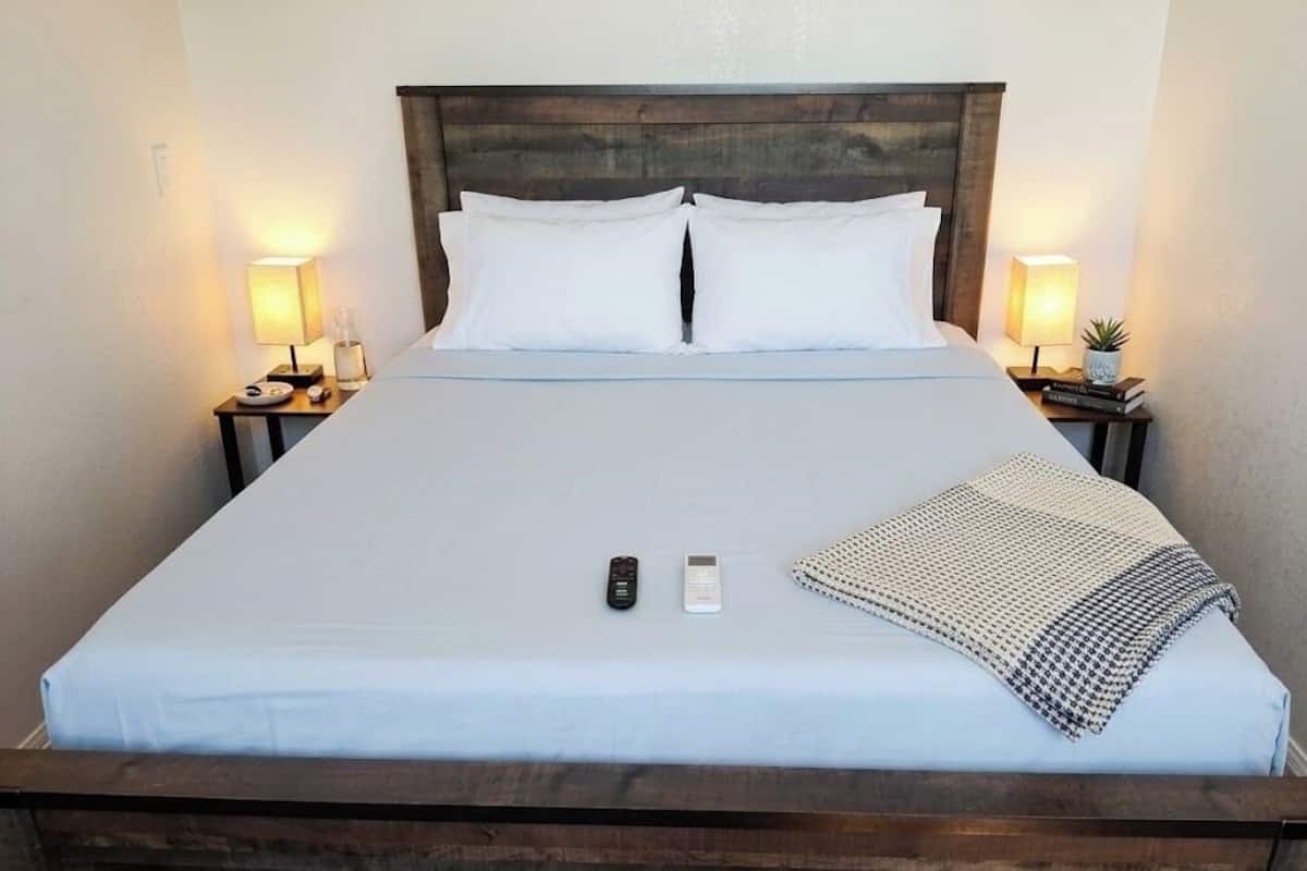 Een net opgemaakt bed met witte lakens, een houten hoofdbord, nachtlampjes, een afstandsbediening, een smartphone en een deken met een patroon.