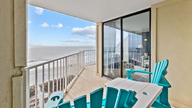 Un balcon avec vue sur l’océan, des chaises turquoise et une porte vitrée donnant sur un espace intérieur.