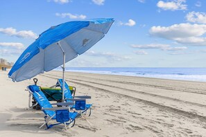 Ein Strand mit einem blauen Sonnenschirm, zwei Liegestühlen und einem Surfbrett.