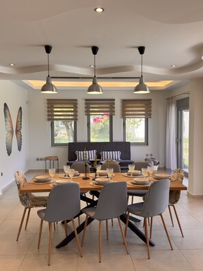 A dining area with a wooden table set for six, surrounded by chairs, under pendant lights, with a view of greenery outside. - Home22 | Sea, Pool & Serenity, Rhodes (Rhodes)