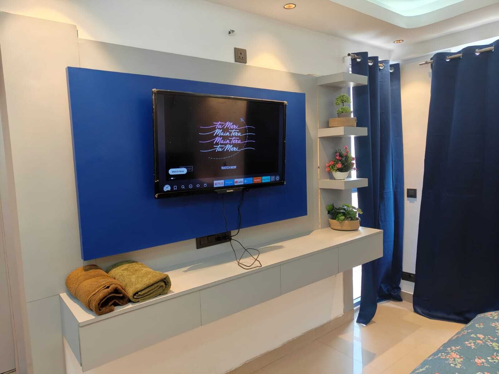 A modern hotel room with a mounted TV, a white console table with towels, a blue accent wall, and a window with curtains.