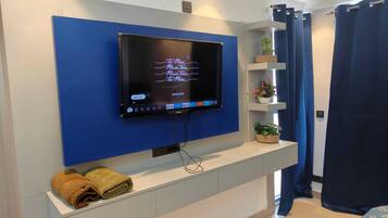 A modern hotel room with a mounted TV, a white console table with towels, a blue accent wall, and a window with curtains.