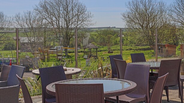 Een terras met rieten stoelen en een glazen tafel, met uitzicht op een tuin en gebouwen in de verte.