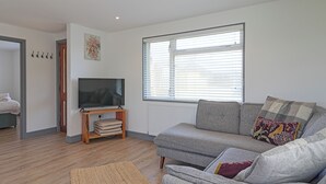 Un salón con un sofá gris, un televisor sobre un mueble de madera, una ventana con persianas y una puerta con perchero.