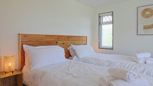 Un dormitorio con dos camas, cabeceros de madera, ropa de cama blanca, una lámpara de noche y una ventana con persianas.