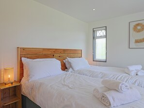 Ein Schlafzimmer mit zwei Betten, einem Holzkopfstück, weißer Bettwäsche, einer Nachttischlampe und einem Fenster mit Jalousien.