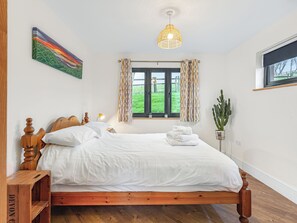 Un dormitorio con cama de madera, ropa de cama blanca, un cuadro, una ventana con cortinas, una lámpara colgante y una planta de cactus.
