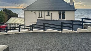 A house with a grey roof, white walls, and a chimney. It has a small window and a door, with a paved area in front. - Bryn Eglwys - UKC9411 (Barmouth)