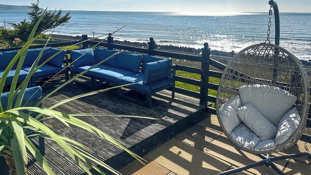A patio with blue outdoor furniture, a hanging chair, and a view of the ocean. - Bryn Eglwys - UKC9411 (Barmouth)