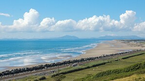 A coastal landscape with a beach, a railway track, and distant mountains under a clear sky. - Bryn Eglwys - UKC9411 (Barmouth)
