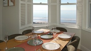 Une table à manger dressée pour six personnes, avec vue sur l’océan grâce à de grandes fenêtres.