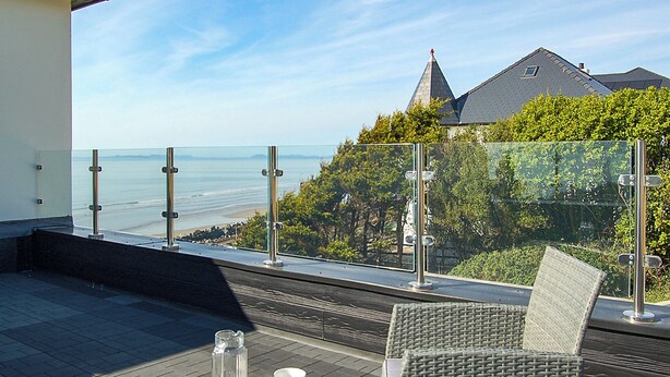 Un balcon doté d'une balustrade en verre donnant sur une plage, une chaise en osier et une table avec deux tasses et une bouteille.