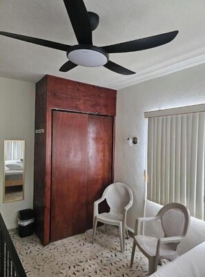 Une pièce avec un ventilateur de plafond, une armoire en bois, une chaise blanche, un lit et une fenêtre avec des stores.