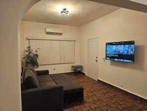 Une pièce comprenant un canapé, un téléviseur fixé au mur, une porte et une fenêtre avec des stores.