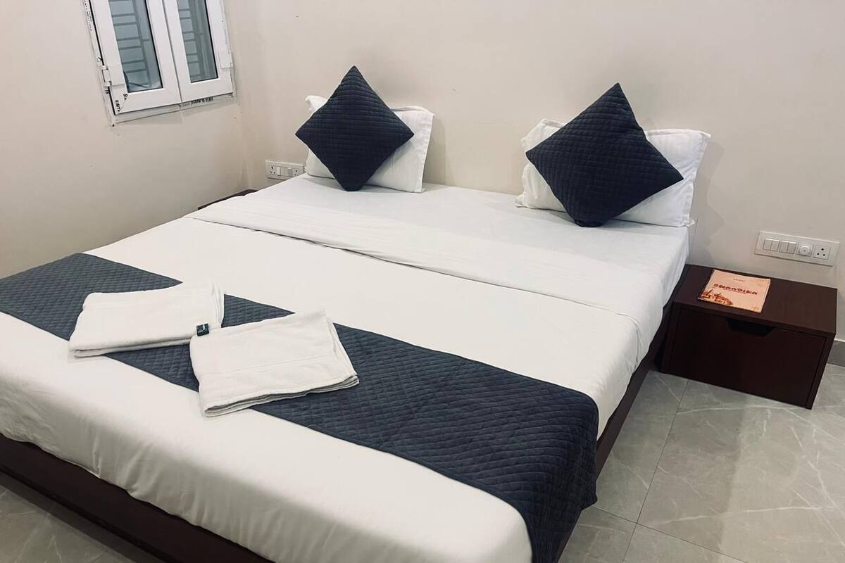A neatly made bed with white and dark blue bedding, a small bedside table with a book, and a window with closed blinds.